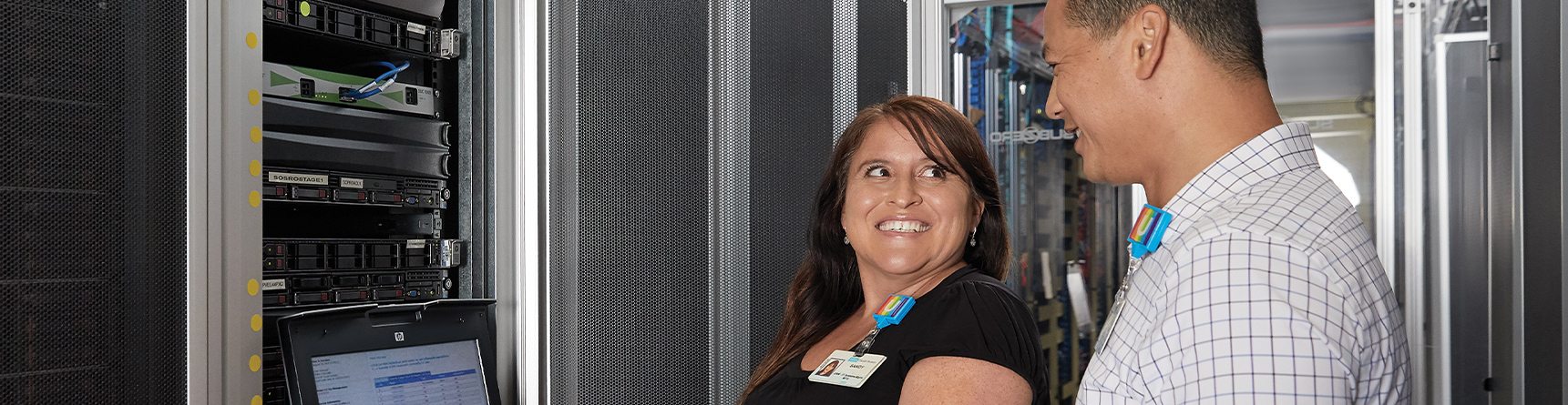Female IT professional and male IT professional smiling while examining computer equipment in large computer server room