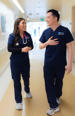 A smiling patient in a hospital bed with a nurse in blue scrubs caring for him, surrounded by medical equipment.