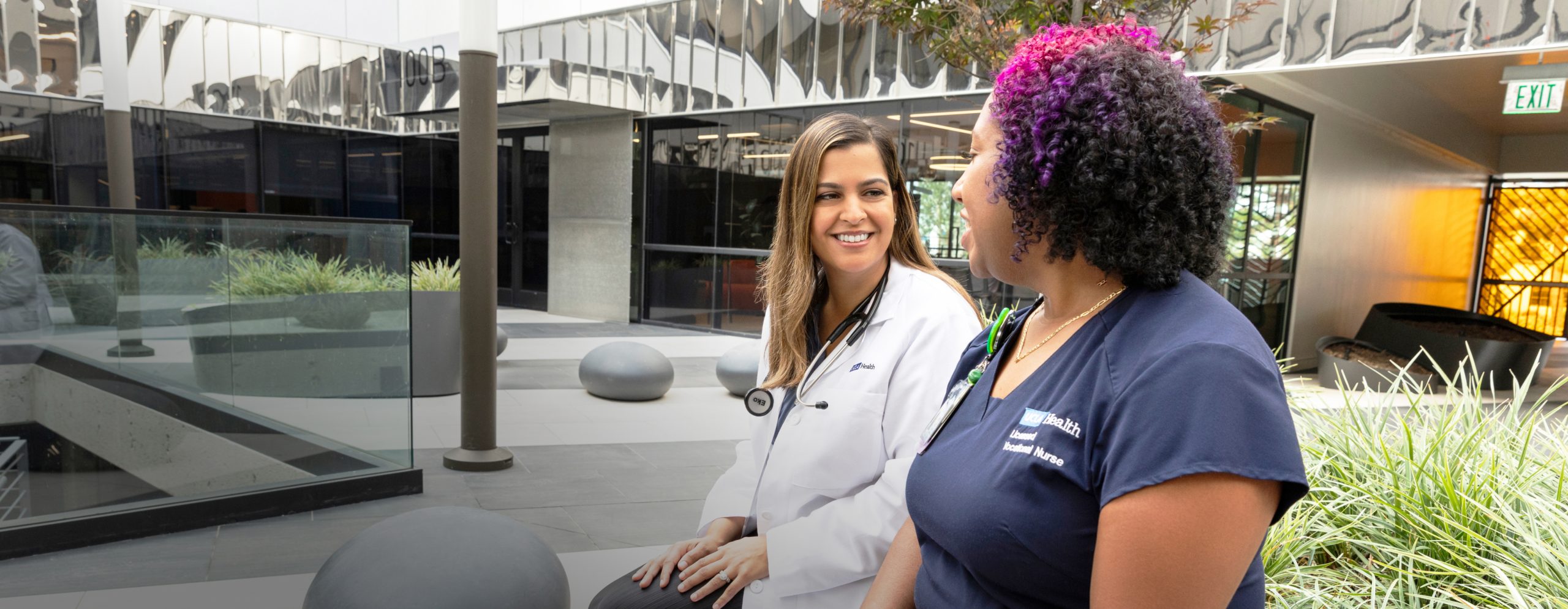 uclahea-cws-who we are A smiling female UCLA Health physician and a smiling female UCLA Health nurse talking together in a courtyard outside of a UCLA Health hospital