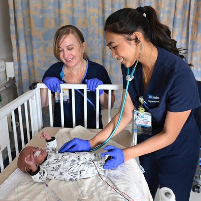Two smiling female nurses in scrubs check a baby in a hospital crib using a stethoscope.
